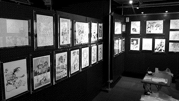 A row of pages from various manga that Hideaki Fujii worked on displayed on two walls of a gallery.
