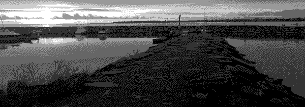 A shale bridge through a body of calm water connected to a long harbor, a mass of land in the distance as a setting sun can be seen through the clouds.